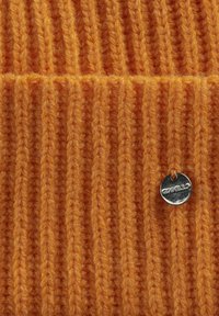 Rippstrick orange Stoff mit strukturierter Oberfläche und einem silbernen Logotag. Das Material wirkt weich und warm.
