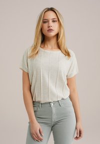 Kreklu ar īsām piedurknēm, bežā krāsā, ar teksturētu dizainu, apaļu kakla izgriezumu un brīvu piegriezumu. Valkāts kopā ar gaiši zaļām, pieguļošām džinsām.