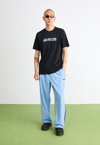 Fekete póló "ADIDAS" grafikával kékszínben, világoskék tréningnadrág fekete oldalcsíkokkal, és fekete klumpák, zöld szőnyegeken állva.
