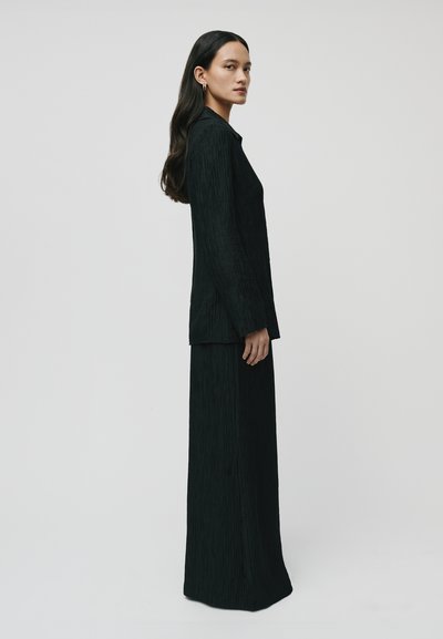 Black two-piece outfit featuring a long, pleated blazer and a matching floor-length skirt. The fabric has a textured finish and a smooth silhouette.