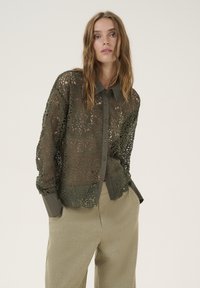 Olivengrøn blondebluse med blomsterdesign og en ensfarvet krave. Knappet foran, lange ærmer og bølget kant. Laget over en lysere top.