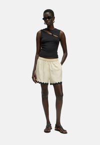Zwart mouwloos topje met zijkanten uitgesneden, crèmekleurige shorts met zwart scalloped rand, en zwarte sandalen, gedragen door een model met kort haar.