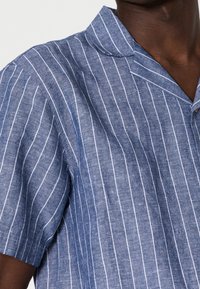 Blauw kortemouwshirt met horizontale witte strepen, gemaakt van textuurstof. Heeft een klassieke kraag en een knoopsluiting.