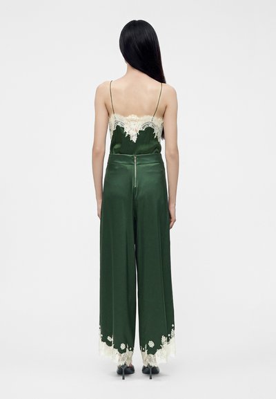 Femme aux longs cheveux noirs portant un pantalon large en satin vert foncé et un caraco assorti avec une bordure en dentelle blanche, vue de dos.