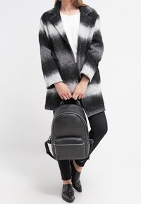 Mochila de cuero negro con forma redondeada, que cuenta con un bolsillo delantero con cremallera y correas ajustables. La lleva puesta una persona con un abrigo en blanco y negro.