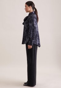 Schwarz leopardgemustertes Blusenshirt mit hohem Kragen, puffigen Ärmeln und Schleifen-Detail. Kombiniert mit schwarzen Jeans im geraden Schnitt und spitzen Schuhen.