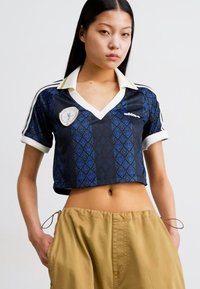 Beskuren marinblå tröja med diamantmönster, vit krage och Adidas-logotyp. Har ett märke på bröstet och korta ärmar.