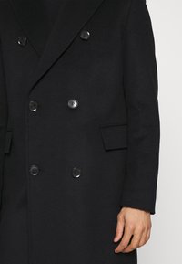 Sort dobbeltradet frakke lavet af uldblanding med store knapper, skarpe revers, frontlommer og en struktureret silhuet.