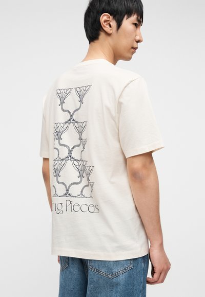 Ανοιχτό μπεζ βαμβακερό μπλουζάκι με μαύρο σχέδιο λουλουδιού σε γραμμή στην πλάτη. Κοντά μανίκια και κλασικό στρογγυλό λαιμό.