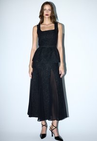 Schwarzes Peplum-Kleid mit einem strukturierten, schimmernden Oberteil, das einen geraden Ausschnitt und durchsichtige Rockpaneele aufweist. Kombiniert mit schwarzen Riemchenschuhen.