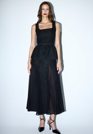 Musta peplum-mekko, jossa on teksturoitu, kimaltava yläosa, suora pääntie ja läpikuultavat hamepaneelit. Yhdistetty mustiin hihnallisiin korkokenkiin.