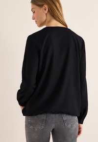 Schwarze Bluse mit glatter Textur, langen Ärmeln und rundem Ausschnitt, kombiniert mit grauen Jeans, die über Gesäßtaschen und eine dezente Waschung verfügen.