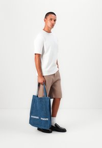 Sac tote en denim de couleur bleue avec des accents cousus, présentant un logo en texte blanc. Un mannequin portant un t-shirt blanc et un short beige se tient devant un fond uni.