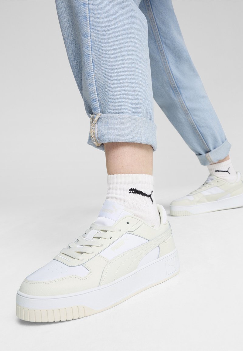 Personne portant un jean bleu clair retroussé, des chaussettes blanches Puma et des baskets blanches Puma à plateforme marchant sur un sol gris.