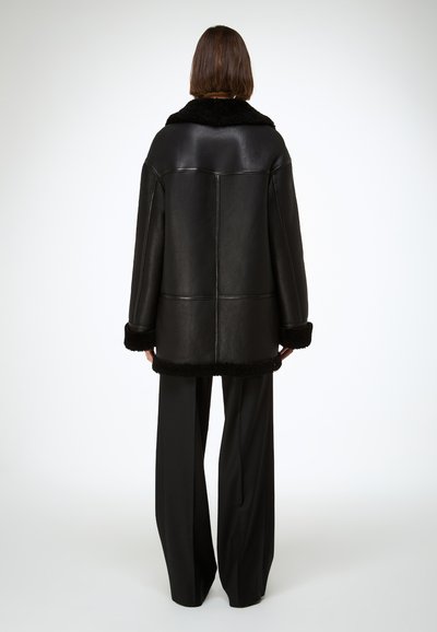 Manteau en cuir noir avec un col et des poignets en peau lainée noire, présentant une forme ajustée et une coupe structurée. Vue arrière montrant un design simple.