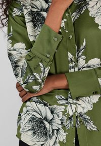 Zelená kvetinová blúzka s bielymi a čiernymi vzormi pivoniek, zapínanie na gombíky, dlhé rukávy s gombíkovými manžetami a hladká textúra látky.