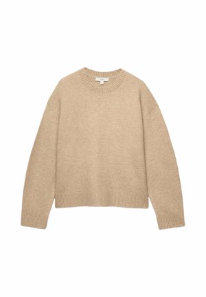 Person mit kurzen lockigen Haaren, die goldene Creolen trägt, einen beigen Langarmpullover und schwarze Hosen, mit dem Rücken zu einem einfarbigen Hintergrund.