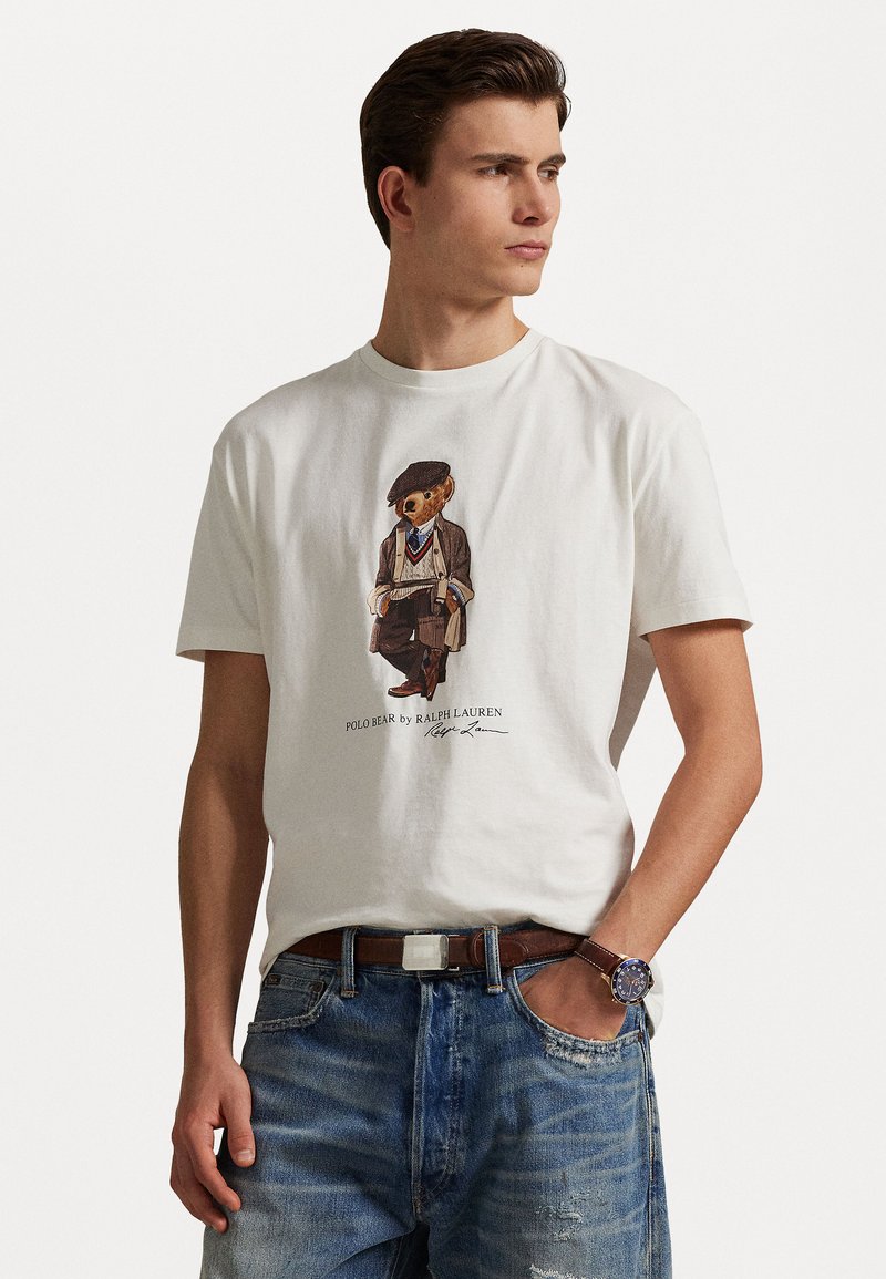 T-shirt en coton blanc avec un motif d'un ours habillé. Accompagné d'un jean en denim bleu, d'une ceinture marron et d'une montre-bracelet. Style décontracté.