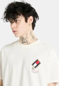 Camiseta color crema con un logo cuadrado que presenta secciones en rojo, blanco y azul. Corte clásico con mangas cortas y una textura suave.