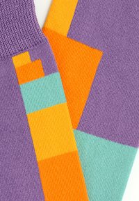 Strikkede sokker med et geometrisk mønster i lilla, orange og teal. Består af blødt stof med en ribbet kant og farveblokke i kontrast.