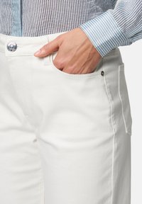 Pantalon en denim blanc avec coupe classique, poches avant, fermeture à bouton métallique et détails de couture subtils. Manchon de chemise rayée visible.