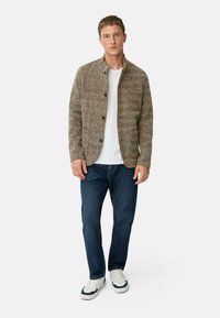 Brauner strukturierter Cardigan mit Stehkragen, Knopfverschluss, zwei vorderen Taschen, getragen über einem weißen T-Shirt und kombiniert mit blauen Jeans und weißen Sneakers.
