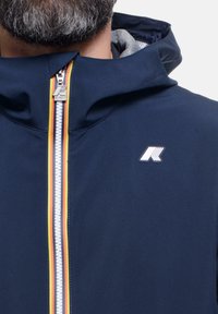 Giacca a vento blu navy con zip frontale, caratterizzata da dettagli rossi, gialli e arancioni; include una toppa con logo sul lato sinistro del petto. Texture liscia.