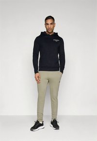 Camisola com capuz preta com logótipo frontal, calças de treino cinza claro de corte justo e sapatos desportivos pretos, num fundo cinzento simples.