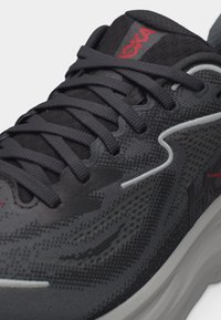 Κοντινή λήψη μαύρου και γκρι αθλητικού παπουτσιού HOKA με υφή υφάσματος, μαύρα κορδόνια, κόκκινο λογότυπο στη γλώσσα και άσπρη μαξιλαρωμένη σόλα.