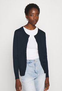 Marineblauwe cardigan van zacht materiaal, met een ronde halslijn, knoopsluiting en lange mouwen, gecombineerd met een wit t-shirt en lichte jeans.