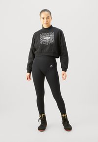 Svart kortermet hettegenser med hvit geometrisk logo, kombinert med svarte tights med en liten hvit logo. Modell som har på seg svarte treningssko.