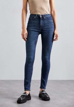 Jean slim bleu foncé porté avec des mocassins noirs épais et un haut beige sans manches sur une personne debout devant un fond gris uni.