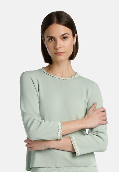 Top mint verde, cu mâneci lungi, cu un finisaj texturat; prezintă un tiv din dantelă la decolteu și la manșete, creând detalii subtile.