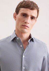 Giovane uomo con capelli castani corti che indossa una camicia a quadri blu e bianca su uno sfondo chiaro e semplice.