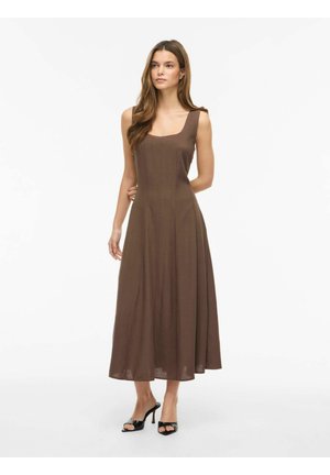 Kvinde stående iført en brun ærmeløs midikjole med firkantet udskæring og sorte åbne tætte højhælede sko på en hvid baggrund.