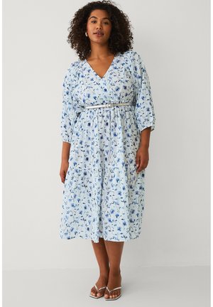 Kvinde iført en lyseblå blomstret midi-kjole med 3/4 ærmer og et tyndt bælte, stående barfodet mod en ensartet hvid baggrund.