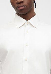Primer plano de una persona que lleva una camisa blanca abotonada con cuello puntiagudo sobre un fondo liso.
