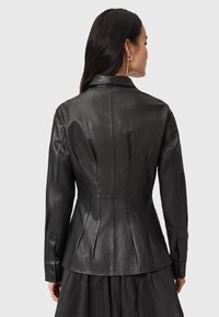 Giacca di pelle nera con una silhouette aderente, spalle imbottite e colletto alto. Presenta polsini con bottoni e cuciture verticali per dare struttura.