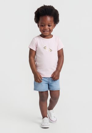 Tout-petit aux cheveux bouclés portant un t-shirt rose clair avec des motifs d’abeilles, un short en jean et des baskets blanches, souriant et avançant.