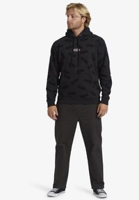 Felpa nera con cappuccio caratterizzata da un sottile motivo logo a tutto campo, cappuccio con coulisse e tasca a marsupio frontale, abbinata a pantaloni scuri e sneakers.