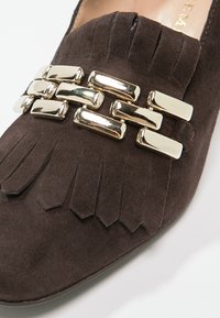 Chaussure en daim marron avec un bout pointu, présentant des détails en frange et une accent en chaîne dorée sur le devant. Texture lisse et semelle plate.