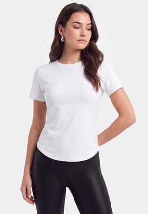 Kvinde med langt mørkt hår bærer en enkel hvid t-shirt med korte ærmer og sorte leggings, kigger ned med den ene hånd på hoften.