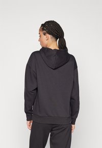 Sudadera negra de tejido suave, con capucha, puños acanalados y un corte relajado. Detalle del logo visible cerca del dobladillo.