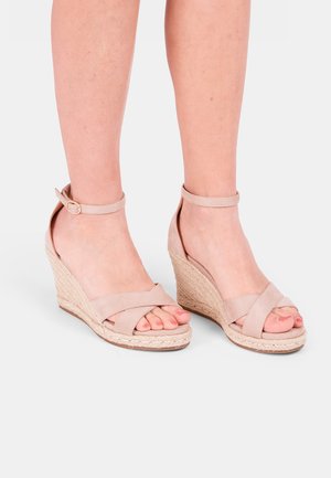 Beige wedge sandals featuring cross-strap design, ankle strap with buckle, and textured woven jute platform. Peep-toe style.