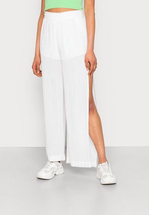 Pantalon blanc léger et fluide avec un tissu texturé et des fentes latérales, porté avec des baskets blanches épaisses. Coupe décontractée avec une taille élastique.