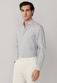 Čovjek obučen u svijetlosivu košulju s tankim prugama i gumbima, uvučenu u hlače visokog struka u boji slonovače, stoji ispred jednobojne pozadine.