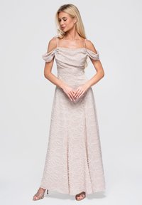Femme blonde portant une robe rose pâle à épaules dénudées avec des motifs circulaires texturés, debout, les mains jointes et regardant vers le bas.