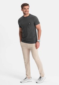 Grå bomulds t-shirt med lomme, cremefarvede slim-fit bukser og hvide mesh sneakers. Enkel, afslappet design med et ur som tilbehør.
