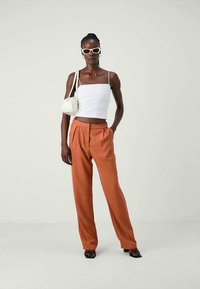 Woman wearing white crop top, rust-colored wide-leg trousers, white shoulder bag, white sunglasses, and black heeled sandals, standing against plain background.