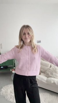 Mlada žena plave kose u ružičastom džemperu s gumbima i crnim hlačama, stoji raširenih ruku u ugodnoj dnevnoj sobi.